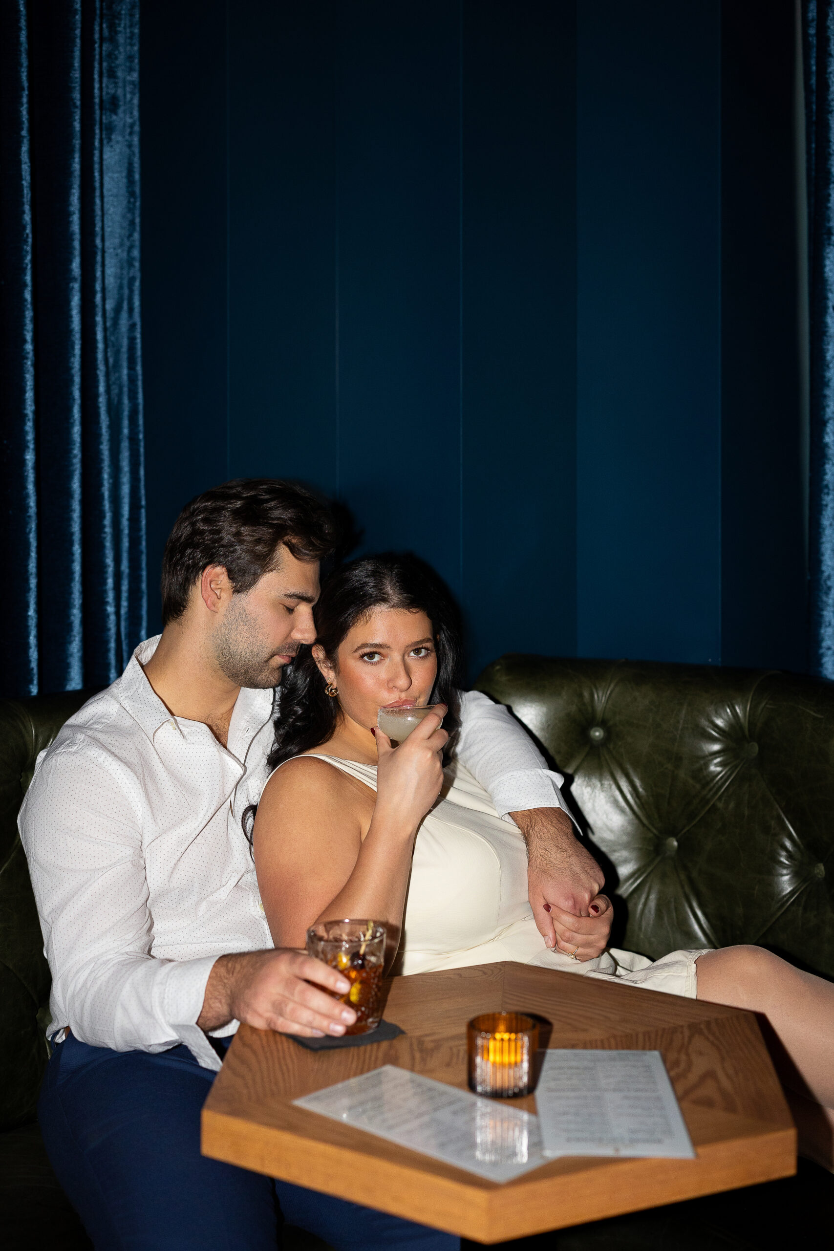 a couple cuddles up in a booth of a dark bar while wearing white and sipping cocktails