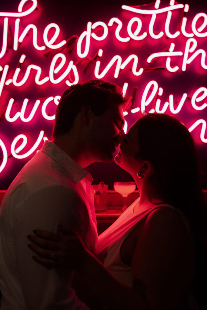 Romantic couple almost kissing in front of pink neon sign at Des Moines bar, engagement photo with moody lighting and cocktails in background.