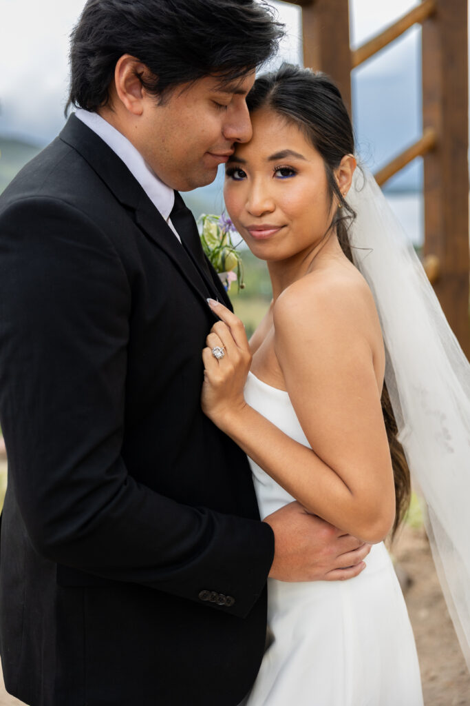 Romantic wedding portrait of bride and groom embracing outdoors, bride wearing strapless gown and veil with engagement ring visible, groom in black suit, photographed at a mountain or countryside venue.
